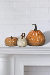 Three ceramic pumpkins on a white fireplace mantle against a white brick wall.