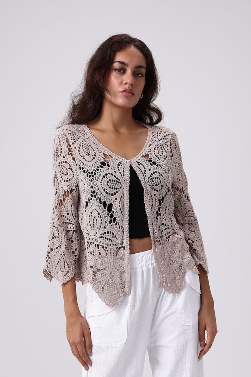 Woman wearing a beige crochet cardigan over a black top and white pants on a light gray background