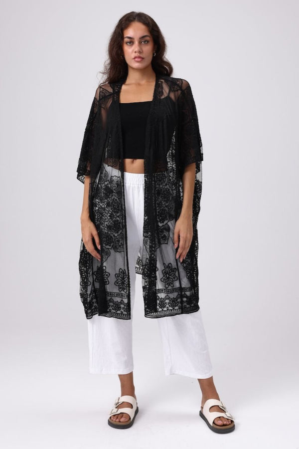 Woman wearing a black lace kimono over a black top and white pants on a white background