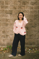 Person wearing a pink cardigan with red cherry patterns against a brick wall.