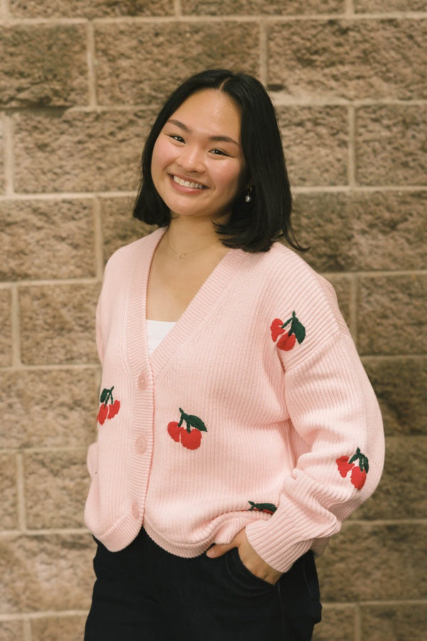 Woman wearing a pink cardigan with cherry designs against a brick wall.