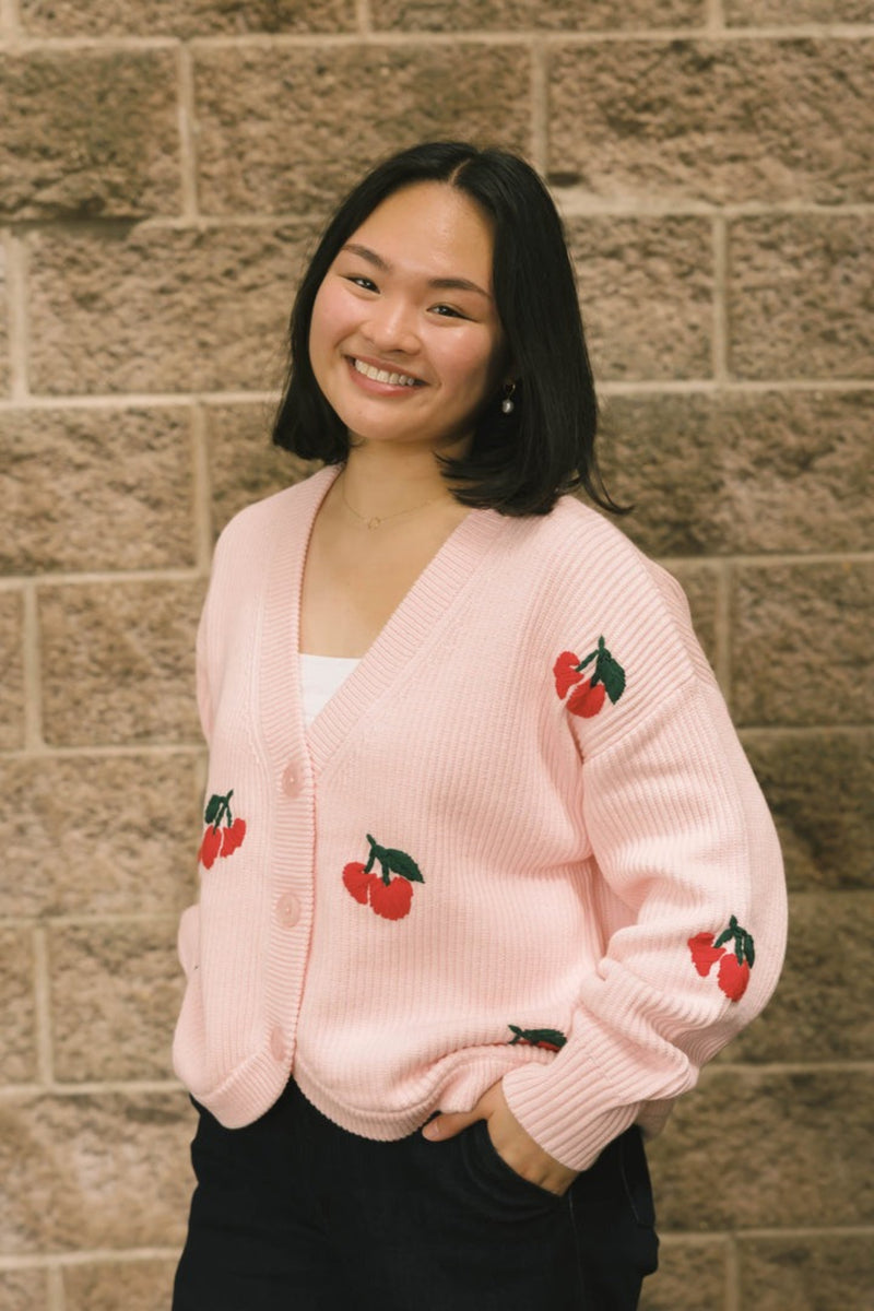 Woman wearing a pink cardigan with cherry designs against a brick wall.