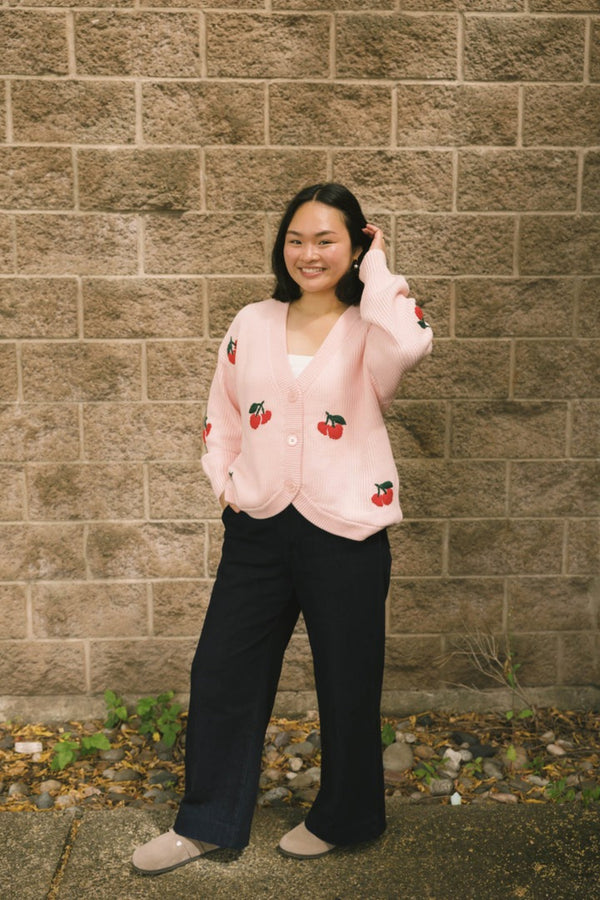 Person wearing a pink cardigan with red cherry patterns against a brick wall.