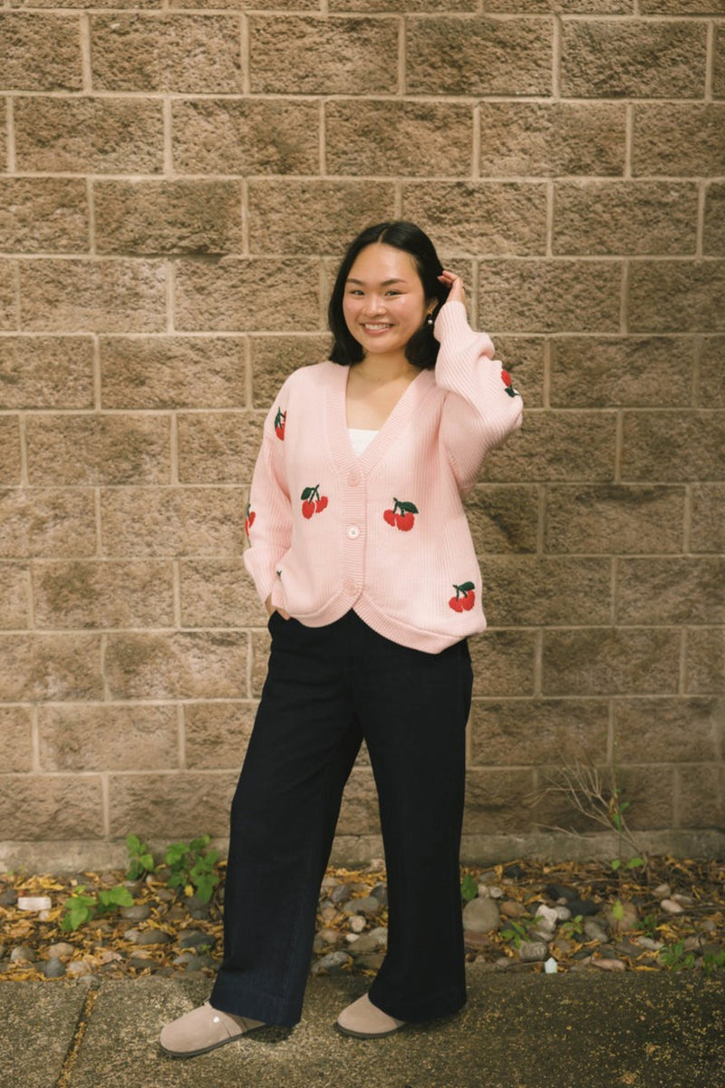Person wearing a pink cardigan with red cherry patterns against a brick wall.