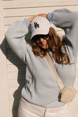 Person wearing a checkered shirt, sunglasses, and a cap with 'LA' logo against a wooden wall.
