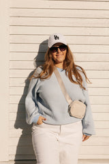 Woman wearing a light blue sweater, white pants, sunglasses, and a white cap with a logo against a beige wall.