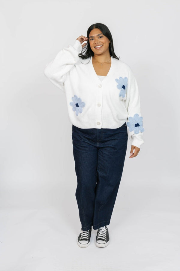 Woman wearing a white cardigan with blue floral patterns and dark pants on a white background
