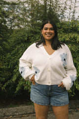 Woman wearing a white cardigan with floral patterns and denim shorts standing outdoors.