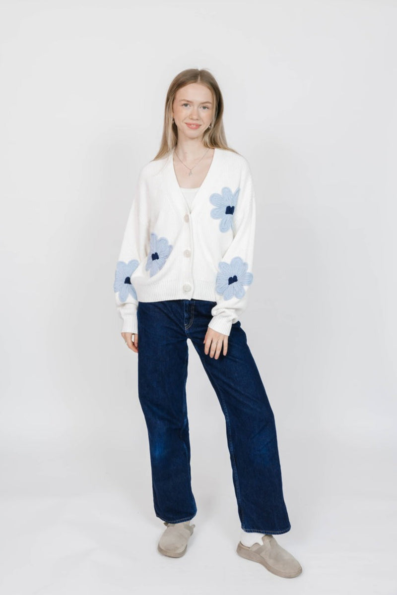 Woman wearing a white cardigan with blue floral patterns and blue jeans on a white background