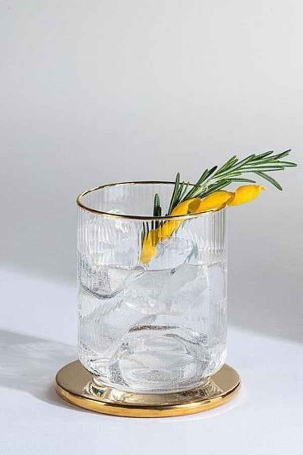 Clear ribbed glass with gold rim filled with ice and a sprig of rosemary on a gold coaster against a light gray background