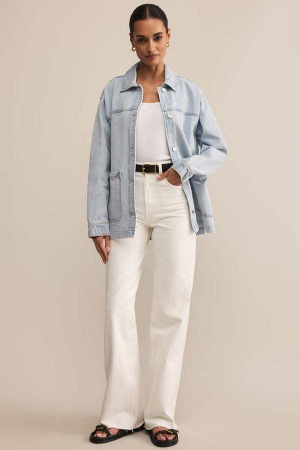 Woman wearing a light blue denim jacket, white top, and white pants on a beige background