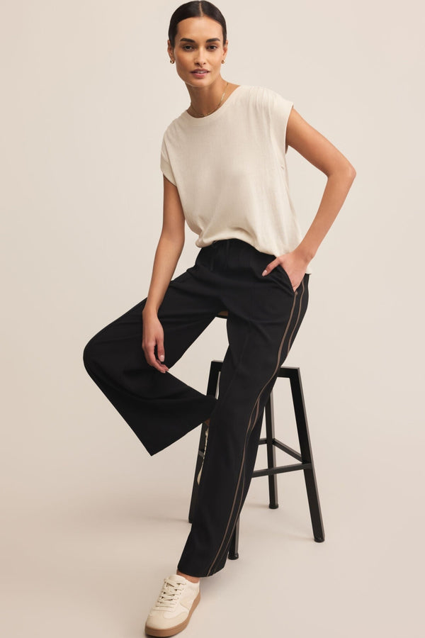 Woman wearing a white top and black pants sitting on a stool against a plain background