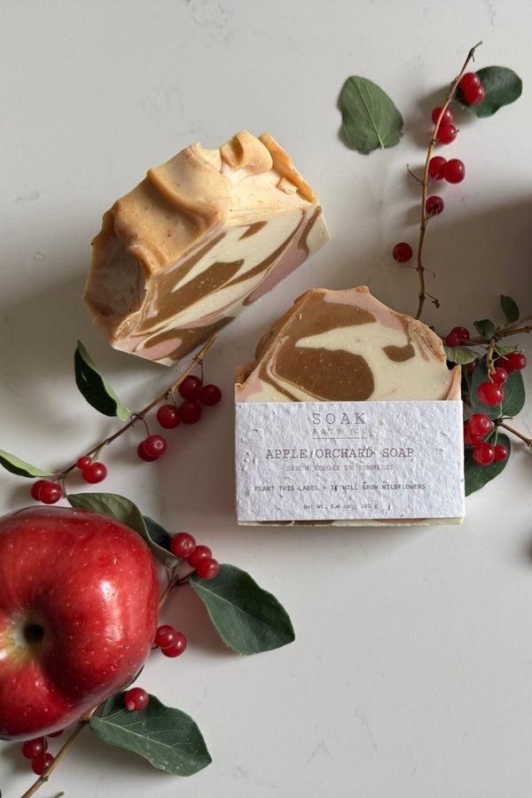 Two pieces of marbled soap with a box, surrounded by red apples and berries on a light surface.