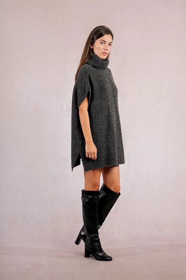 Woman wearing a gray knitted poncho and black knee-high boots on a plain background