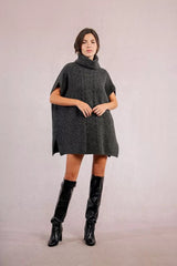 Woman wearing a dark gray knitted dress with a turtleneck and black knee-high boots on a plain background