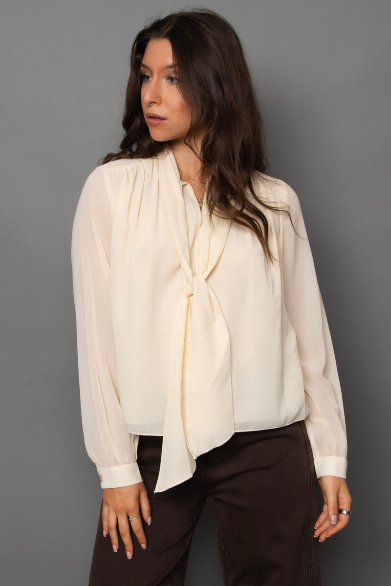 Woman wearing a cream blouse with a tie detail against a gray background