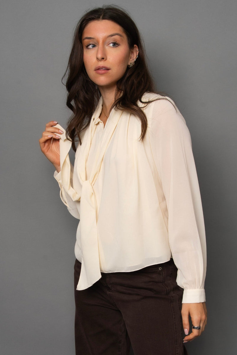 Woman wearing a cream blouse and dark pants against a gray background